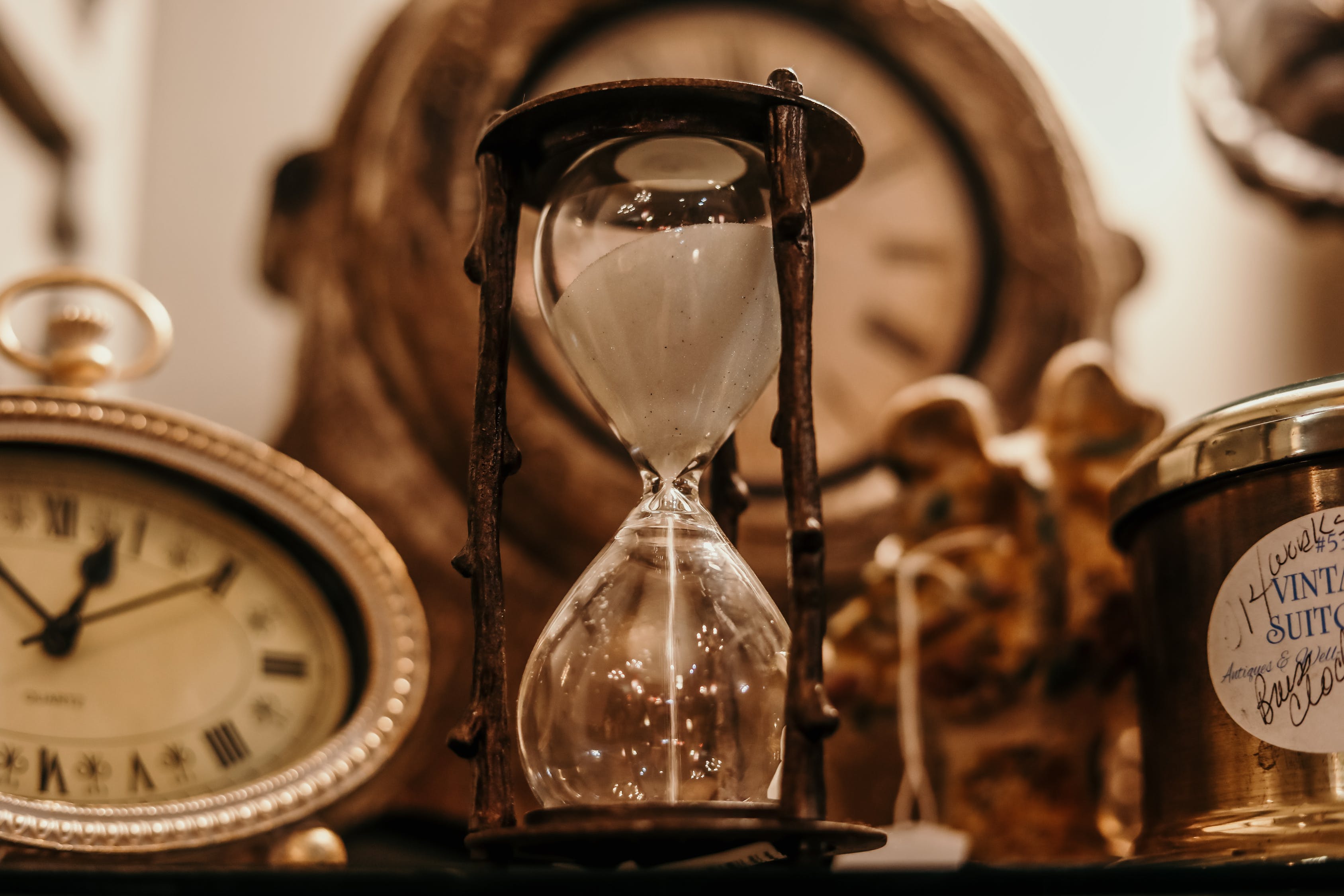  An hourglass timer sits in the foreground with a clock and other objects in the background, symbolizing the need for patience and perseverance in overcoming procrastination.